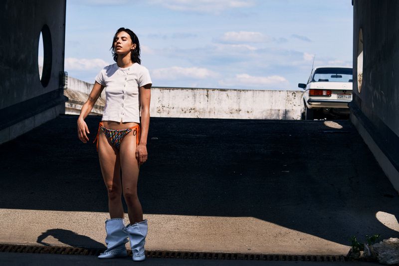 Woman in white top and bikini bottoms standing outside tunnel on sunny day near car. Fashion editorial photo.