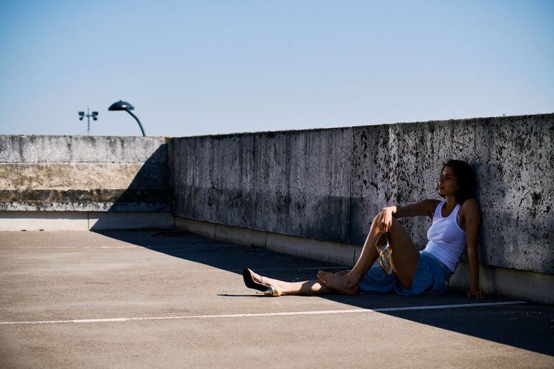 Woman in casual summer wear sits on a concrete surface, leaning against a weathered wall under bright sunlight in an urban setting.