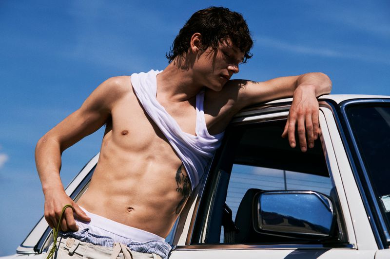 A shirtless young man in a rolled-up white tank top leans against the roof and window frame of a white car under a bright blue sky.
