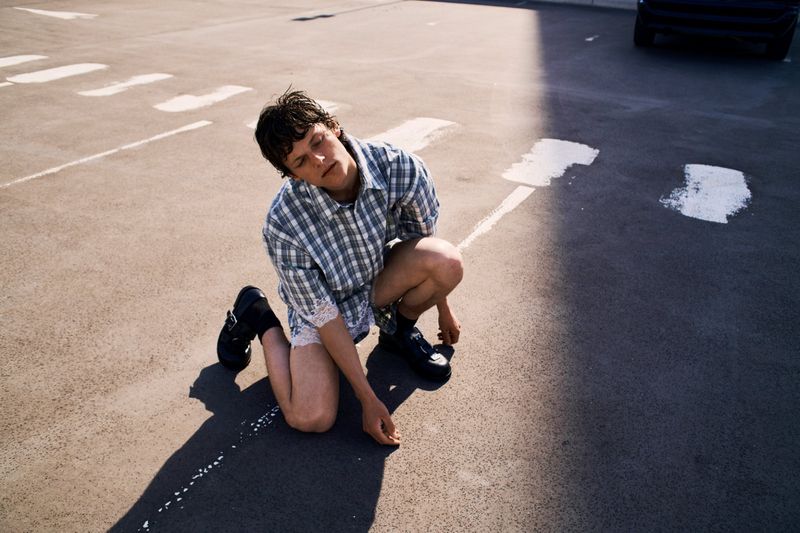 Person wearing a plaid shirt kneels on the pavement of a parking lot, eyes closed, sunlight casting a shadow.