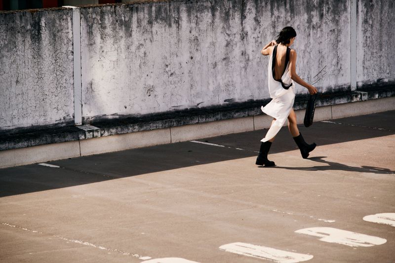 Model walking in an open-air parking structure wearing a white backless dress and black boots for SSENSE project HEAT.
