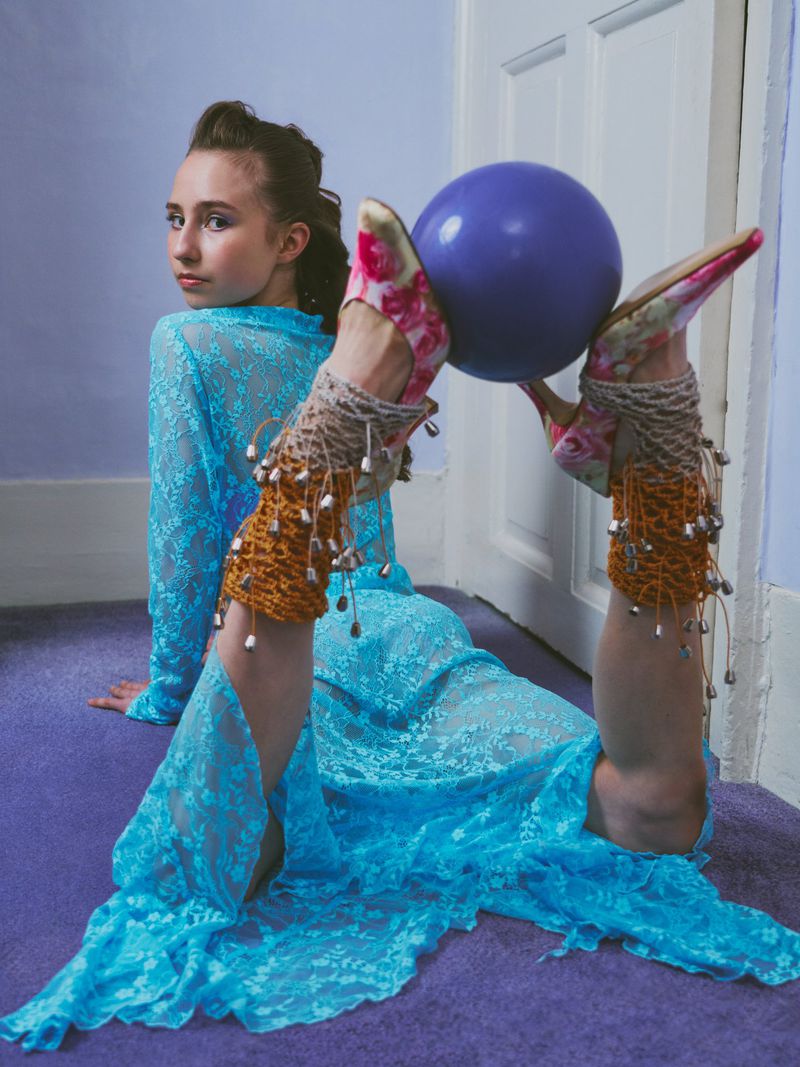 Model in a bright blue lace dress lying on a purple carpet holding a purple sphere between floral patterned high heels.