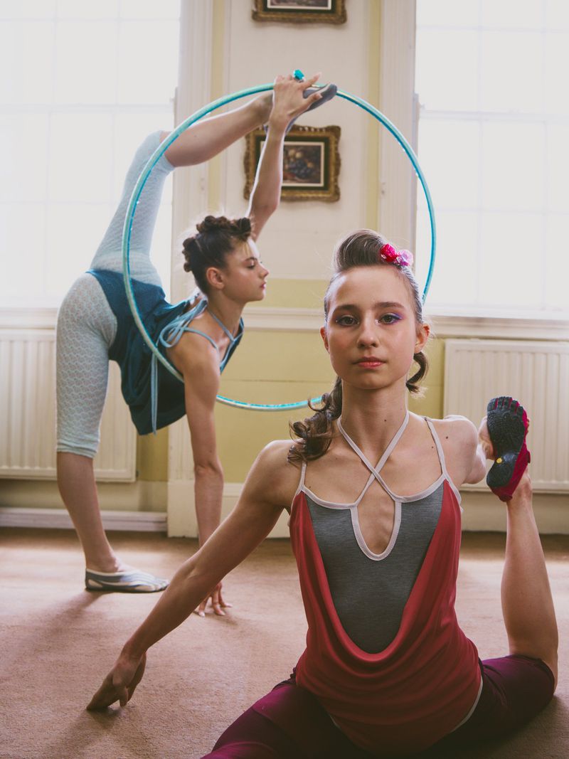 Two young female gymnasts holding flexible poses with a turquoise hula hoop inside a naturally lit interior room.