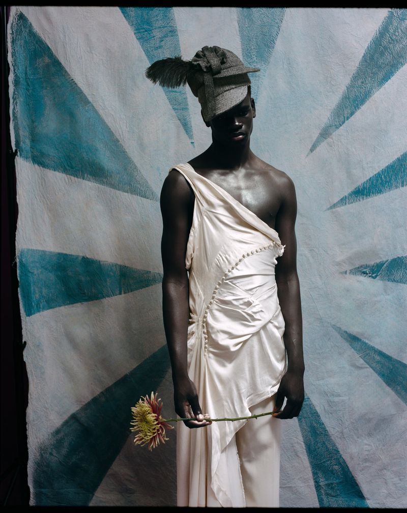 A Black male model wearing a draped cream gown and a grey feathered headpiece holding a yellow flower against a blue and white sunburst backdrop