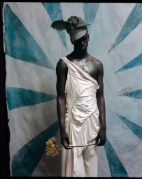 Black male model in an ivory one-shoulder draped garment and feathered hat holding a flower against a painted sunburst backdrop.