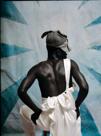 Back view of a Black man wearing a grey tweed cap and draped white fabric against a painted blue and white geometric backdrop.