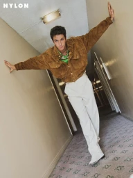Role Model wearing a brown suede jacket and white trousers while posing with arms outstretched against hallway walls.