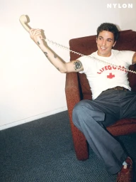 Role Model sitting in an armchair holding a corded telephone handset, wearing a white Lifeguard t-shirt and grey trousers.
