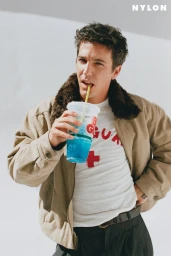 Musician Role Model sipping a blue slushy while wearing a tan shearling-collar jacket and white graphic t-shirt.
