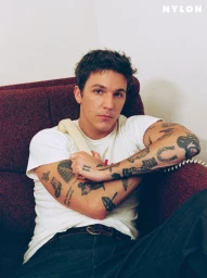 Role Model with visible arm tattoos sitting in a maroon armchair wearing a white t-shirt and jeans for Nylon Guys.