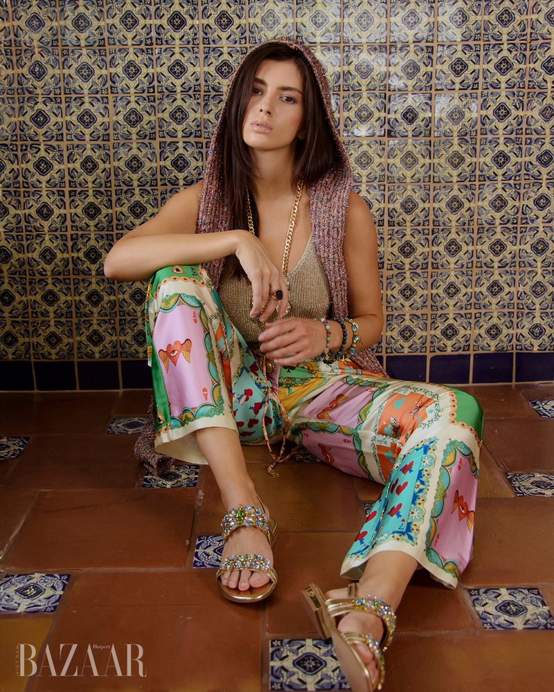 A woman sits on a tiled floor wearing a hooded knit top, silk patterned pants, and jeweled sandals, captured for Harper's Bazaar Vietnam.