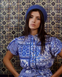 Model in a blue and white patterned dress and blue beret posing in front of a blue decorative tile wall.