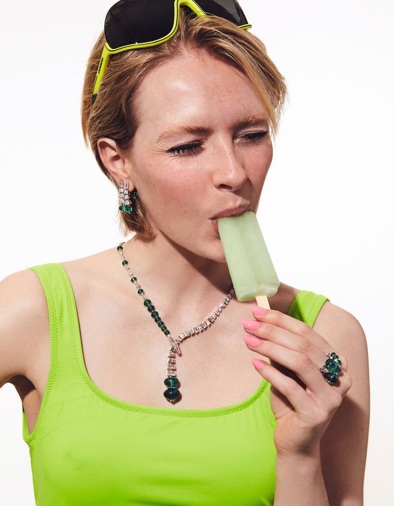 Model in neon green tank top eating a green popsicle wearing sunglasses and gemstone jewelry against a white background