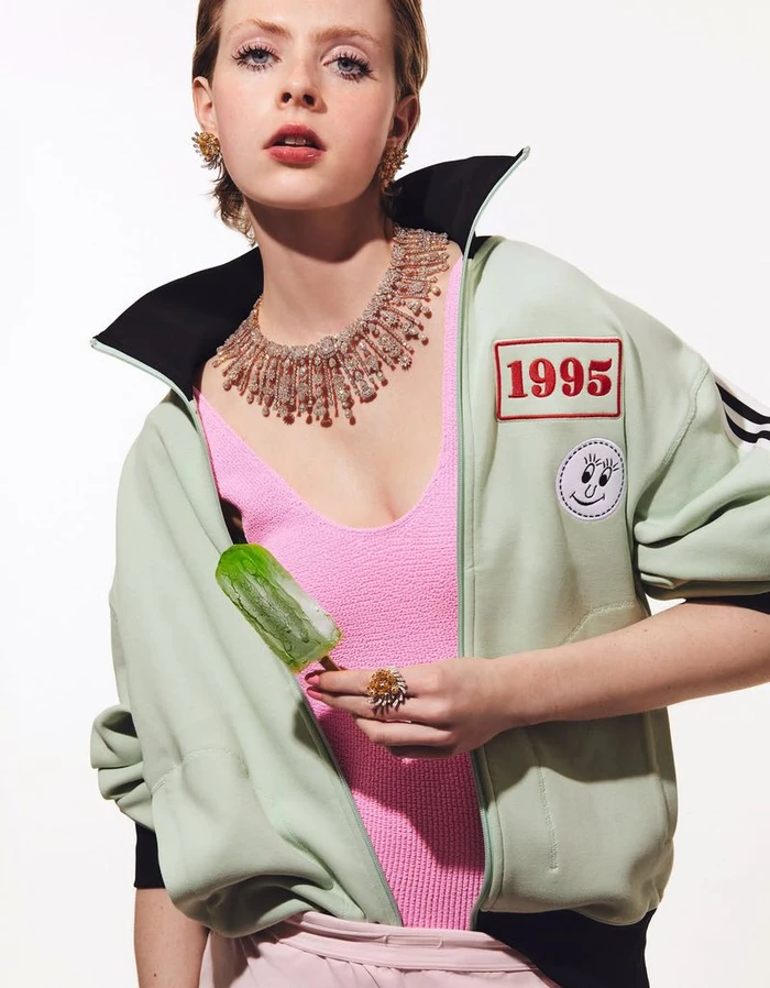 Model Em Verleun wearing a mint track jacket, pink top, and diamond fringe necklace while holding a green popsicle against a white backdrop.