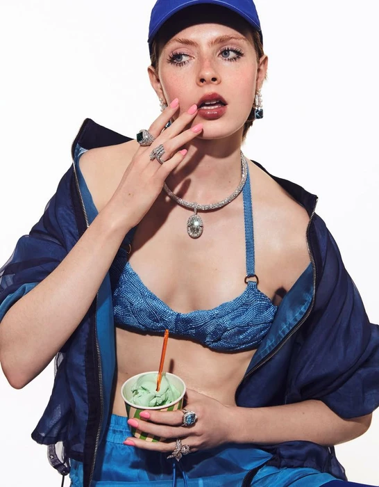 Model Em Verleun in a blue cap, blue bikini top, fine jewelry, and navy jacket holding a cup of mint gelato on a white seamless backdrop.