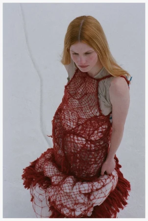 Model Lainey Hearn wearing an open-weave crimson mesh garment with fringe against a pale background for Snow Drift.