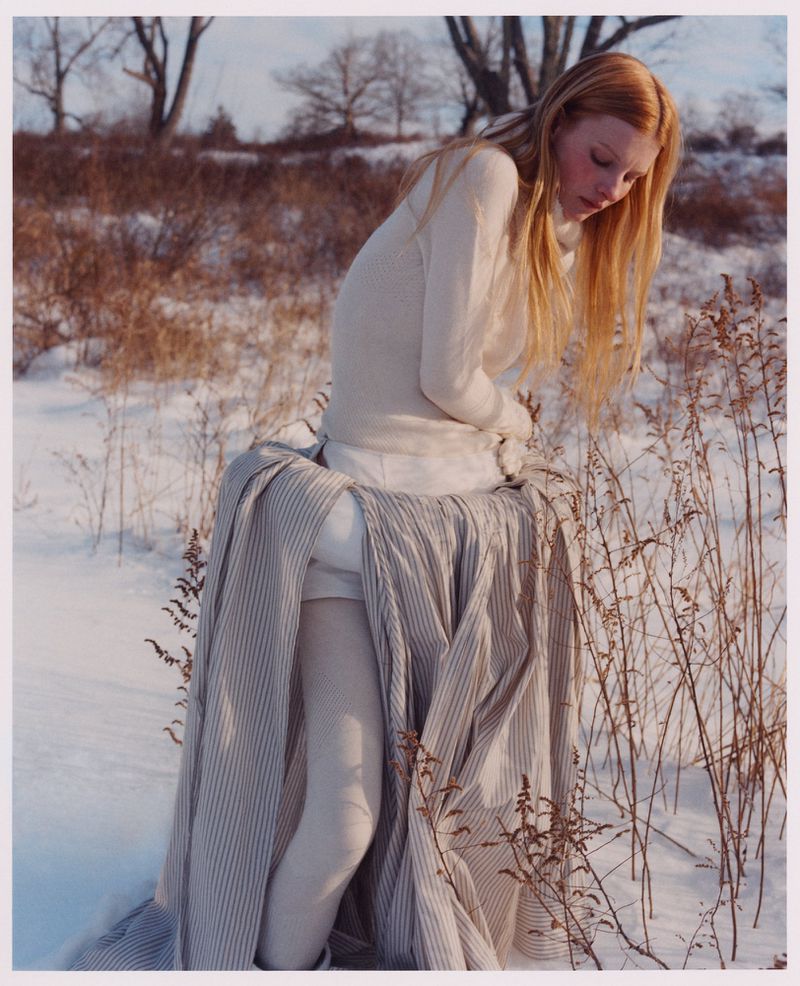 Model with long blonde hair wearing a white sweater and voluminous skirt standing outdoors in a snowy landscape.