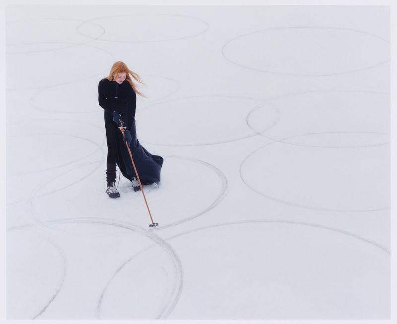 Model Lainey Hearn standing in an all-black ensemble holding a ski pole on a snowfield etched with large geometric circles.
