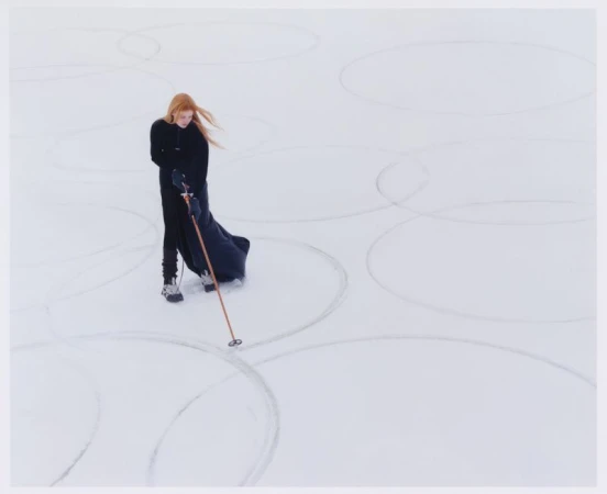 Model Lainey Hearn standing in an all-black ensemble holding a ski pole on a snowfield etched with large geometric circles.