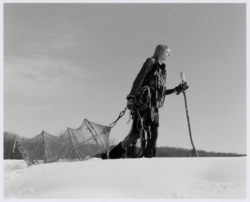 Model wearing dark textured outerwear pulling a netted sled across a snowfield holding a staff