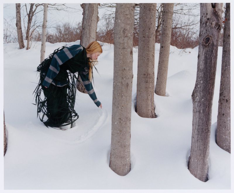 Model Lainey Hearn wearing a structured black sculptural garment leaning into deep snow among tree trunks in a winter forest.
