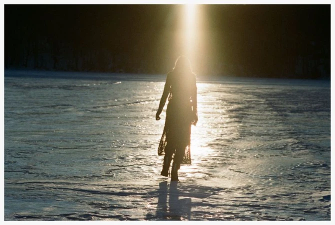 Silhouette of a model walking across a sunlit frozen snowfield under a bright vertical flare of natural backlight.