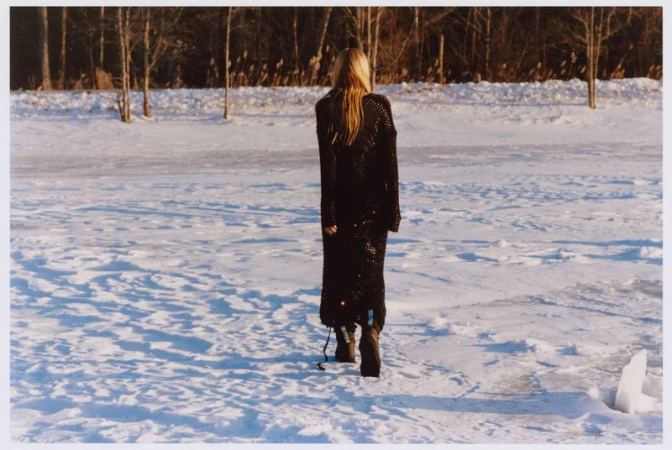 A blonde woman in a long black knit dress walks away across a snow-covered landscape towards a distant line of bare winter trees.