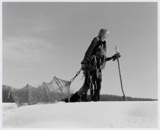 Model wearing dark textured outerwear pulling a netted sled across a snowfield holding a staff
