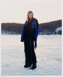 Model Lainey Hearn standing in a snowfield wearing a blue and black windbreaker jacket and black knit dress.
