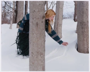 Model Lainey Hearn in a striped sweater and fringe skirt touches deep snow among bare tree trunks in a winter woodland.