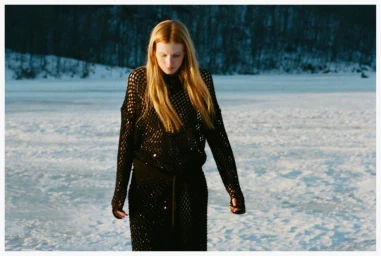 Model Lainey Hearn wearing a black open-knit dress outdoors in a snow-covered winter landscape with distant snowy trees.
