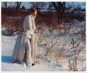 Model wearing a cream knit turtleneck and sculptural draped fabric structure in a snow-covered field with bare trees.