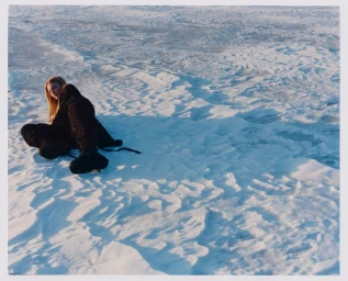 Model Lainey Hearn in a black winter outfit seated on a vast textured snowfield during daylight.