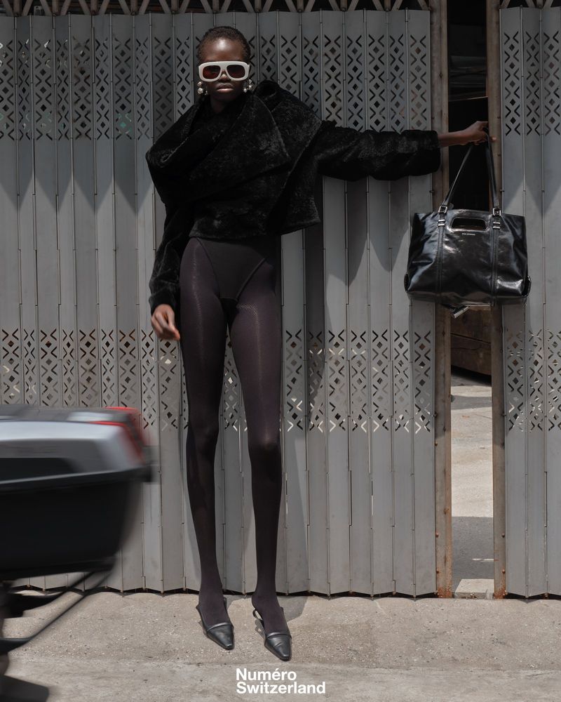 Model Akolde Meen wearing an all-black outfit, white sunglasses, and pointed heels against a grey metal folding gate.