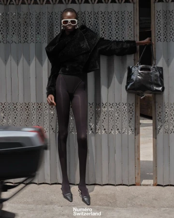 Model Akolde Meen wearing an all-black outfit, white sunglasses, and pointed heels against a grey metal folding gate.