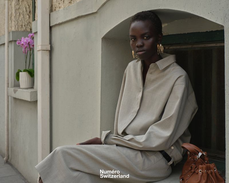 Model Akolde Meen wearing a beige relaxed shirt and gold earrings, seated in an arched exterior window alcove with a brown leather bag.