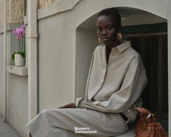 Model Akolde Meen wearing a beige relaxed shirt and gold earrings, seated in an arched exterior window alcove with a brown leather bag.