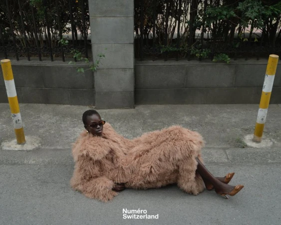 Model Akolde Meen lying on a concrete street wearing an oversized beige shaggy textured coat and dark sunglasses.
