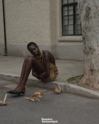 Model Akolde Meen crouching on a street curb in a gold satin backless dress and black kitten heels for Numéro Switzerland.