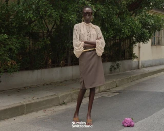 Akolde Meen in a sheer beige blouse and taupe pencil skirt stands on a street with dark sunglasses next to a purple cauliflower.