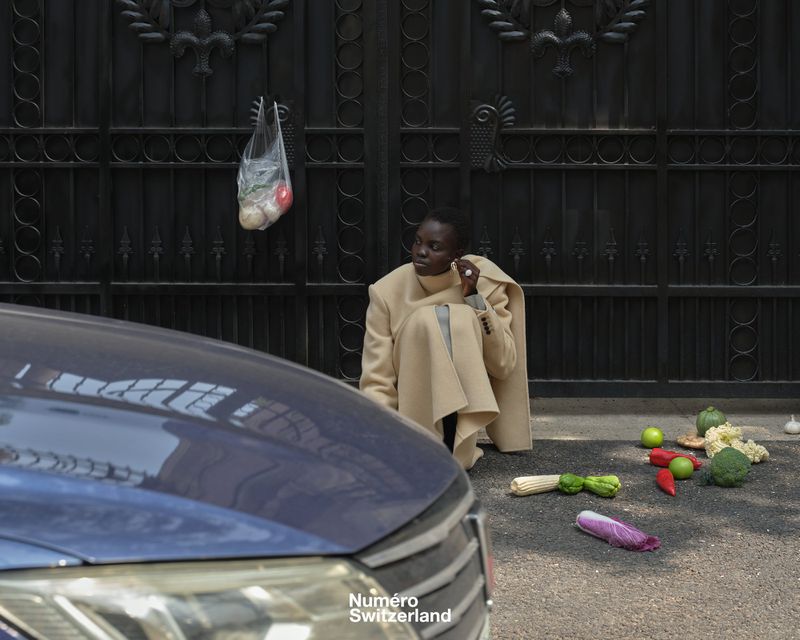 Model Akolde Meen crouching in a beige tailored coat next to scattered vegetables on asphalt before a black wrought-iron gate.