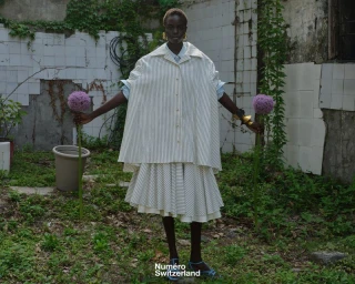 Model Akolde Meen wearing an oversized striped cape shirt and pleated skirt holding two large purple allium flowers in an overgrown yard.