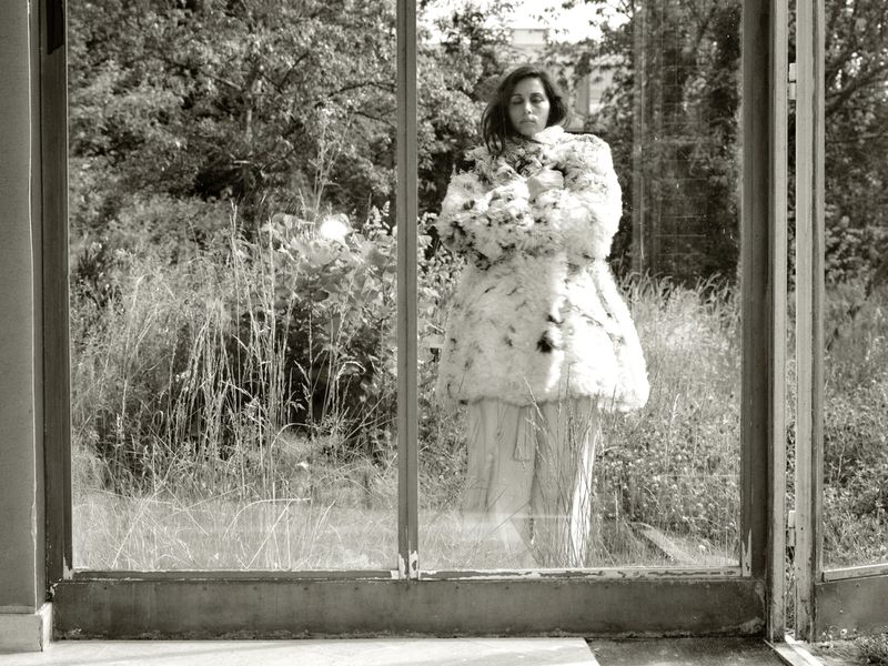 Yasmine Hamdan standing behind glass window frames wearing an oversized fuzzy patterned coat in front of overgrown garden greenery.