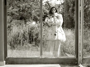Yasmine Hamdan standing behind glass window frames wearing an oversized fuzzy patterned coat in front of overgrown garden greenery.