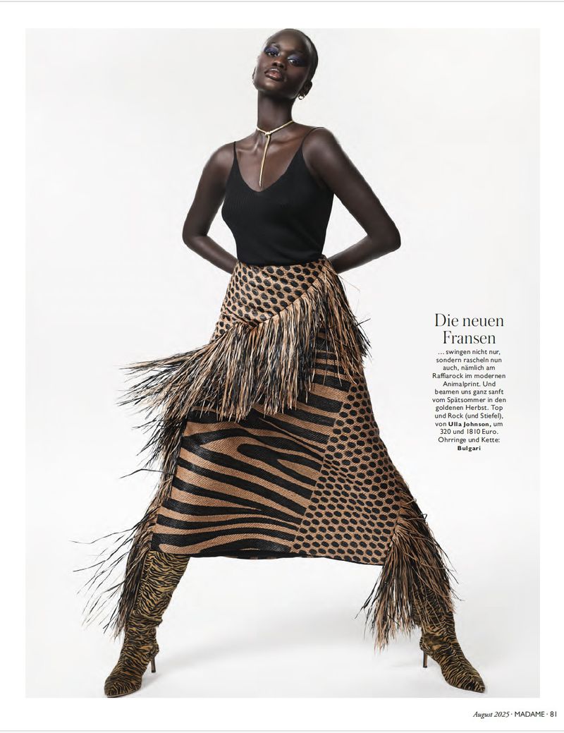 Fashion editorial shot of a tall black woman with bold makeup, wearing a black top and animal-print skirt with fringe, posing against a white backdrop.