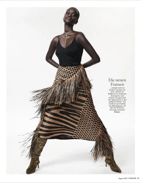 Fashion editorial shot of a tall black woman with bold makeup, wearing a black top and animal-print skirt with fringe, posing against a white backdrop.