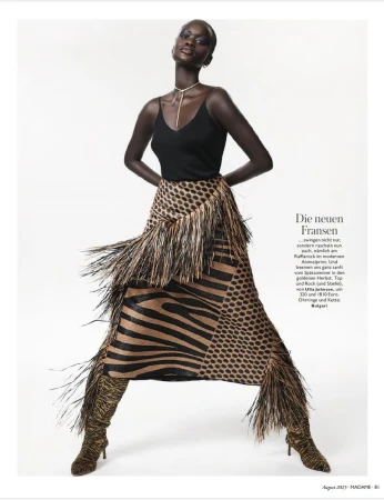 Fashion editorial shot of a tall black woman with bold makeup, wearing a black top and animal-print skirt with fringe, posing against a white backdrop.