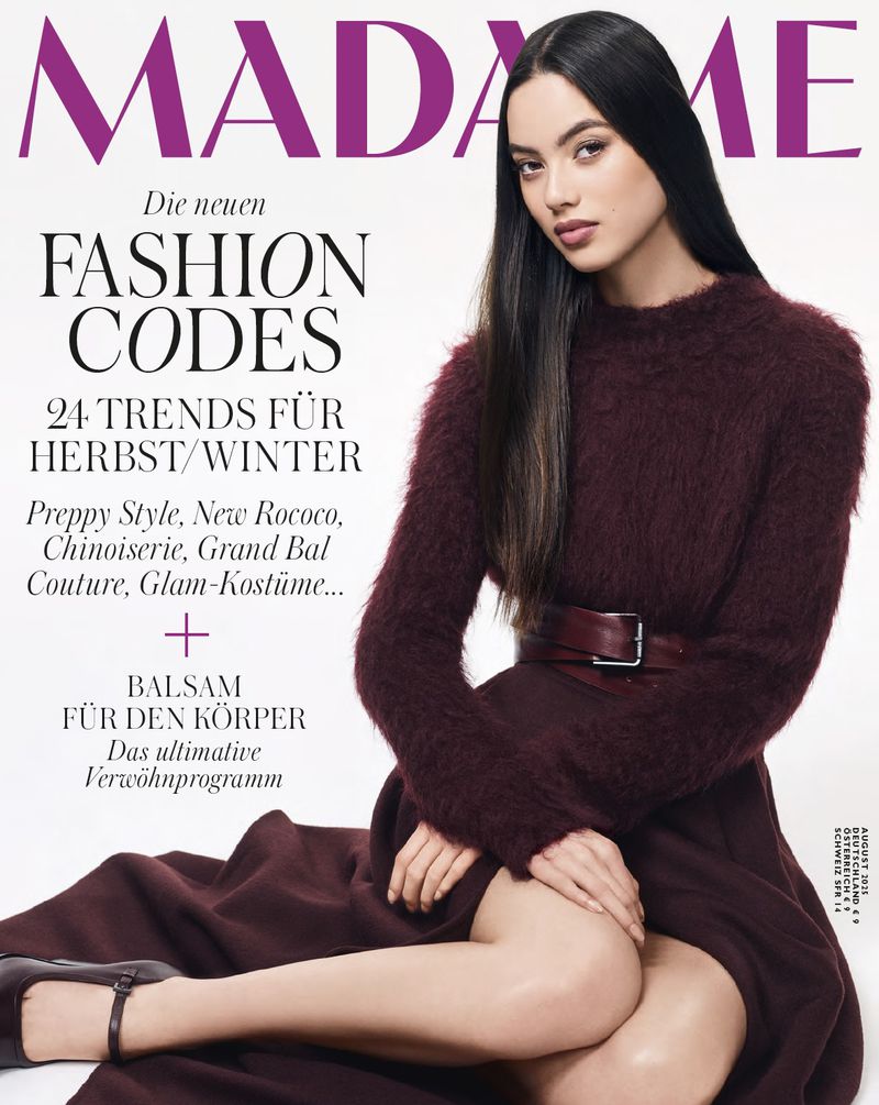 Magazine cover featuring a brunette woman in a maroon dress and belt, seated with her legs crossed against a white background.
