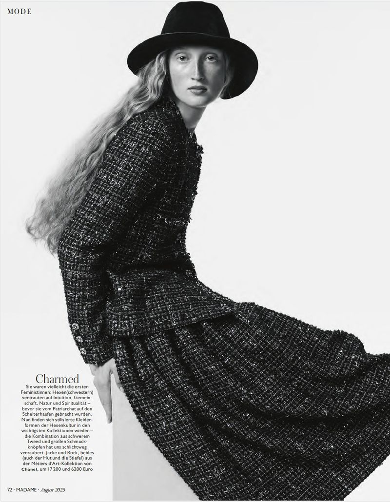 Fashion model wearing a black wide-brim fedora and a textured dark tweed jacket with matching skirt against a white studio background.