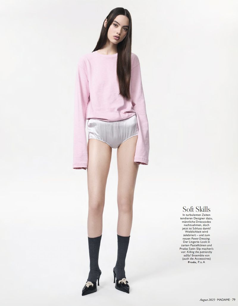Full-length shot of a young woman in a pink sweater and silver shorts, styled with dark socks and high heels against a white backdrop.